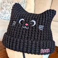 Black knitted cat hat with facial features including white small whiskers, inverted eyes that point towards a small, round pink nose, a small smile, and the beanie shaped into ears and 'boop' text on a mannequin.