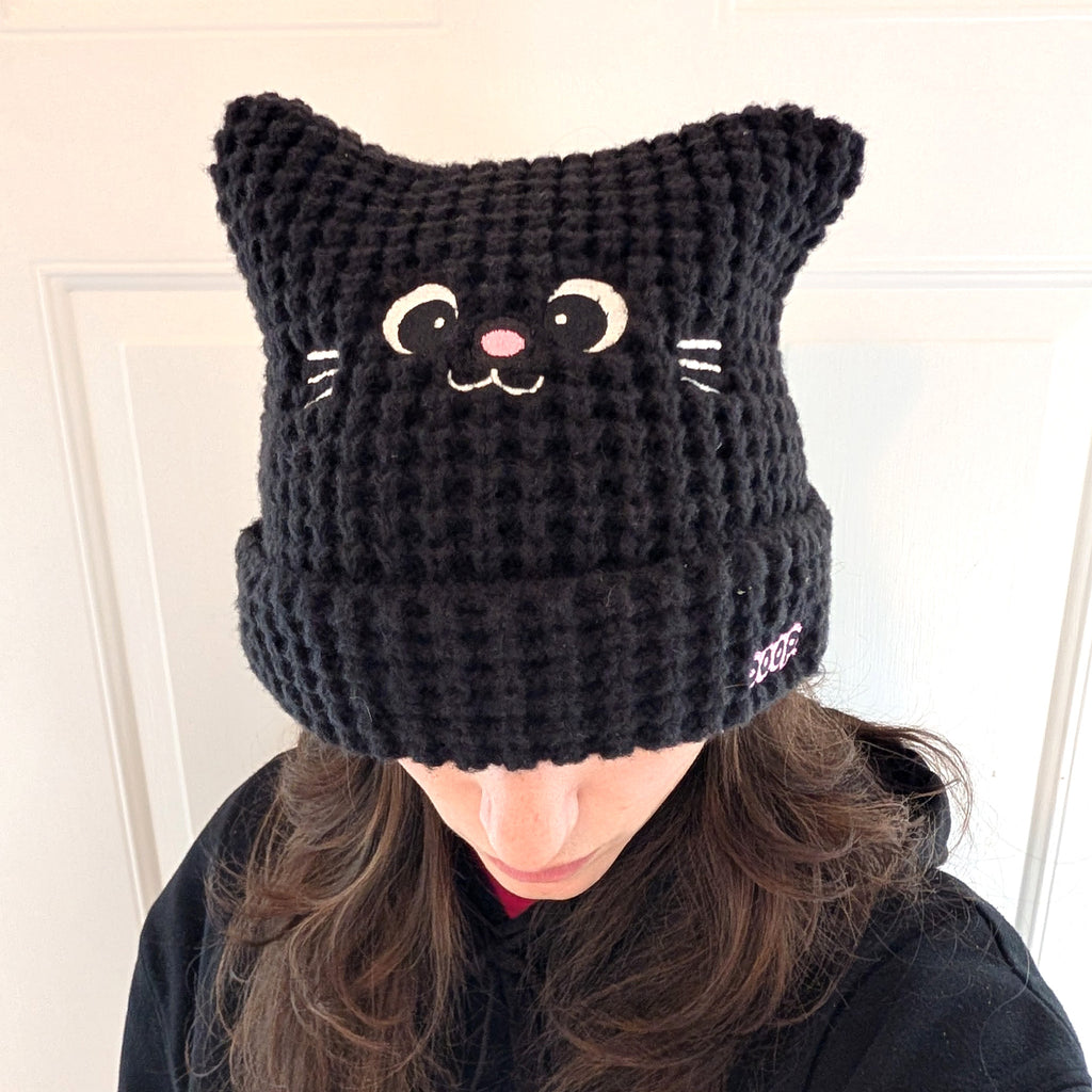 Black knit cat hat with facial features worn by a person indoors.