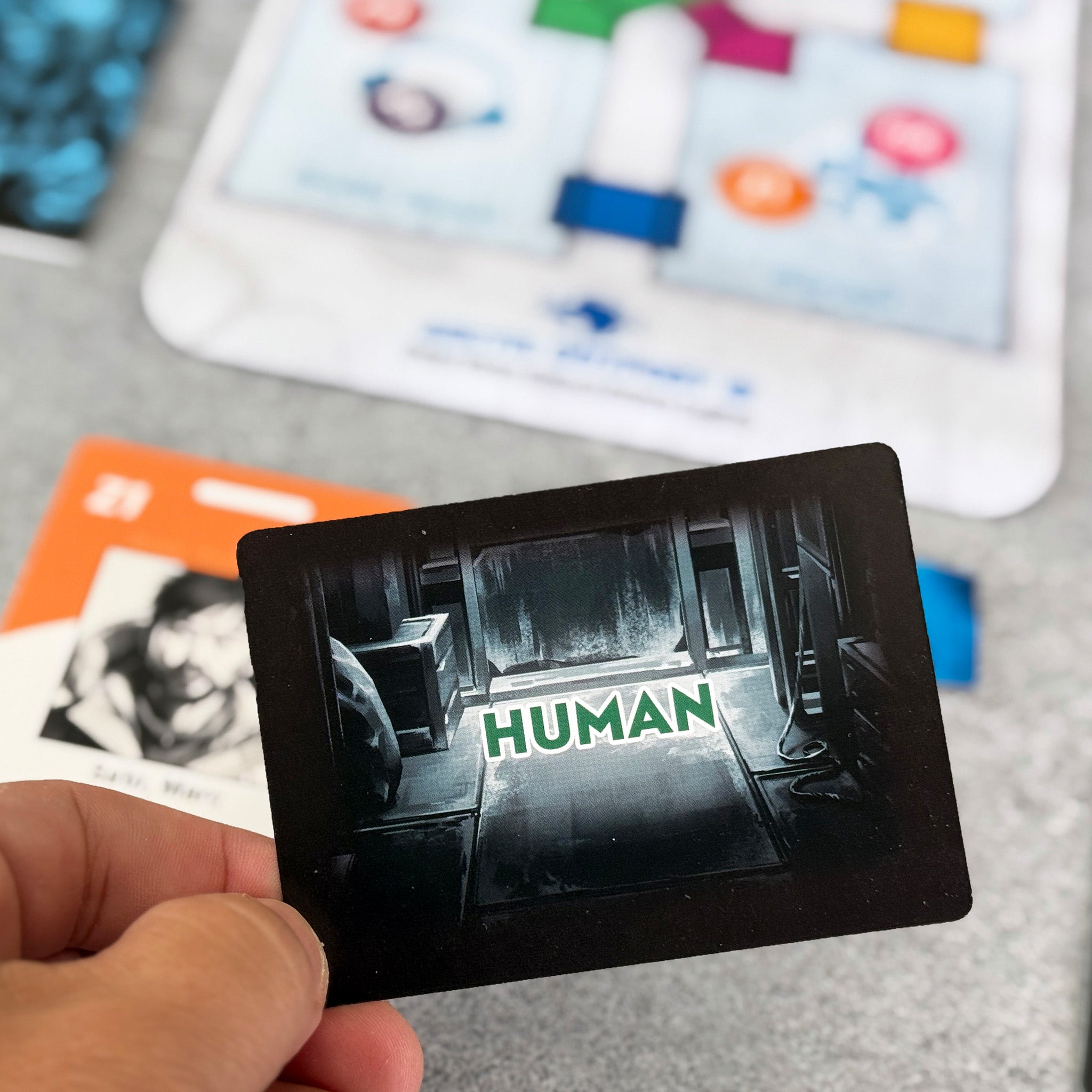 Card with 'HUMAN' text held by a hand, with a colorful board game in the background.