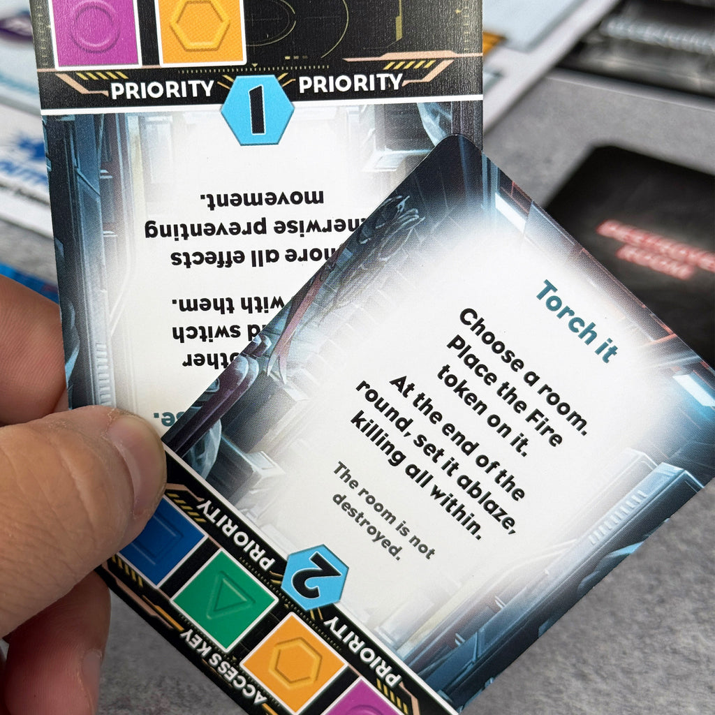 Close-up of a card with game text and priority markers held by a hand. The text reads: Torch it, choose a room, place the fire token on it. At the end of the round, set it ablaze, killing all within. The room is not destroyed. 